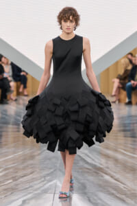 Model walking the runway in a black sleeveless dress with a sculptural, geometric ruffled skirt comprised of overlapping rectangular panels.