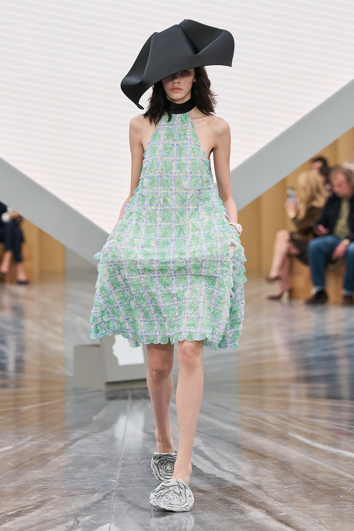 Model on a runway wearing a pastel green and blue halter dress with layered ruffles, an oversized black sculptural hat, and gray shoes.