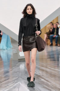 Runway model walks in a black satin blouse with a bow, a brown leather mini skirt, and a large brown shoulder bag.