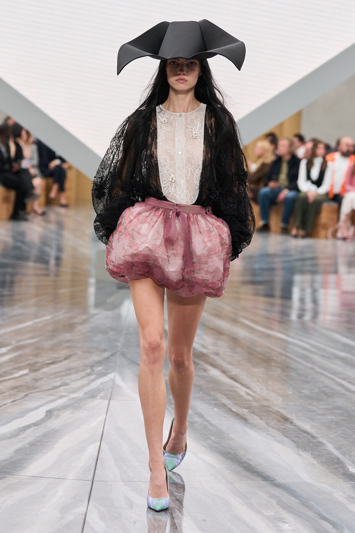 Model on a runway wearing a large black sculptural hat, sheer lace blouse, black lace cape, pink bubble skirt, and iridescent heels.
