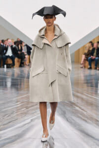 Model walking a fashion show in a pale oversized coat with a large collar and front pockets, paired with a dramatic black sculptural hat and glittery heels, on a glossy runway.