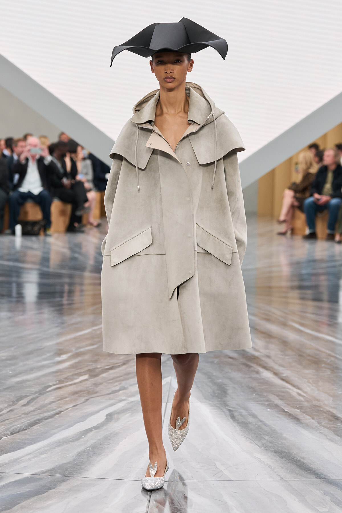 Model walking a fashion show in a pale oversized coat with a large collar and front pockets, paired with a dramatic black sculptural hat and glittery heels, on a glossy runway.