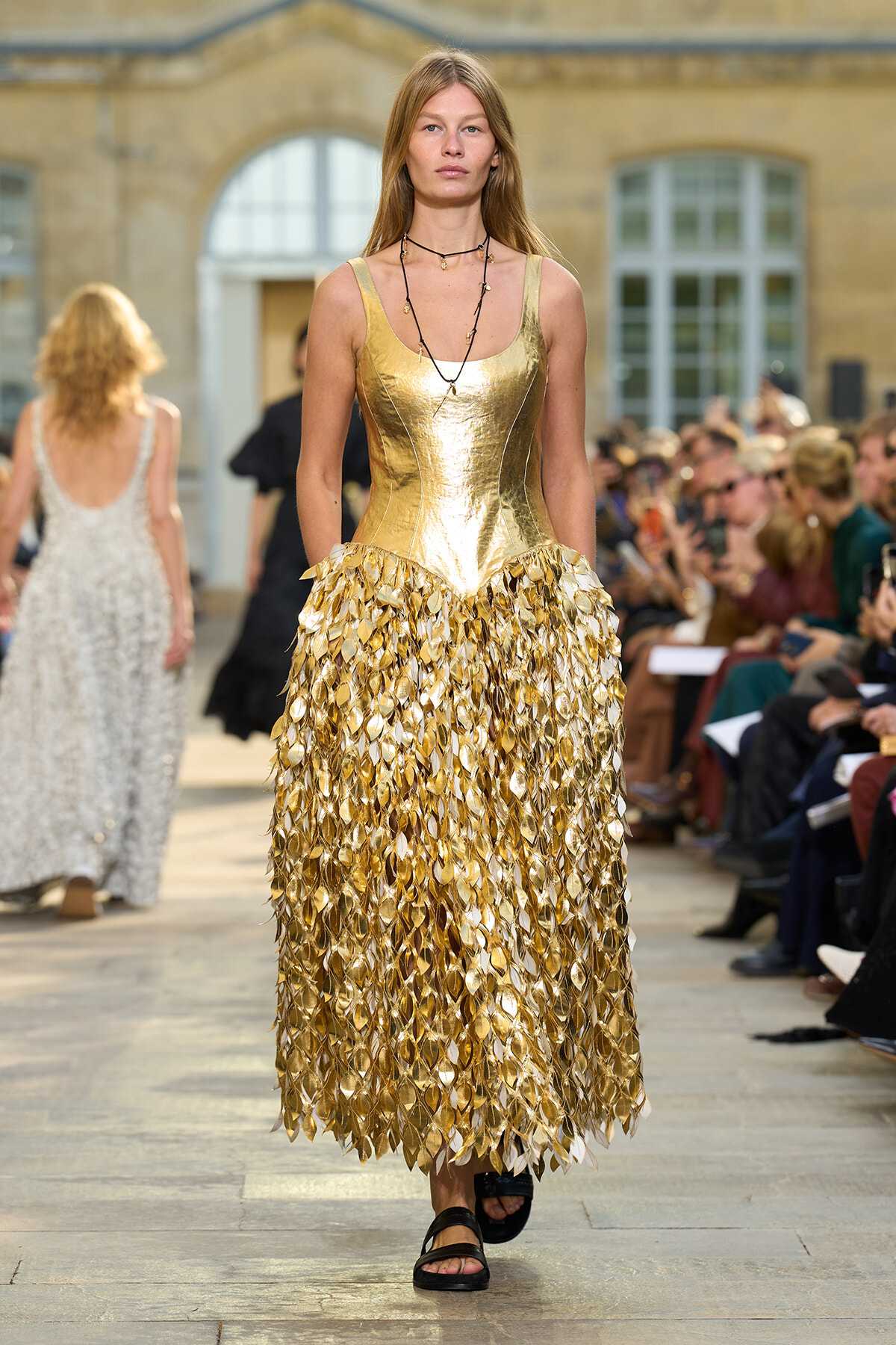 Model walks the runway in a gold metallic dress with a leaf-shaped fringed skirt at a fashion show.