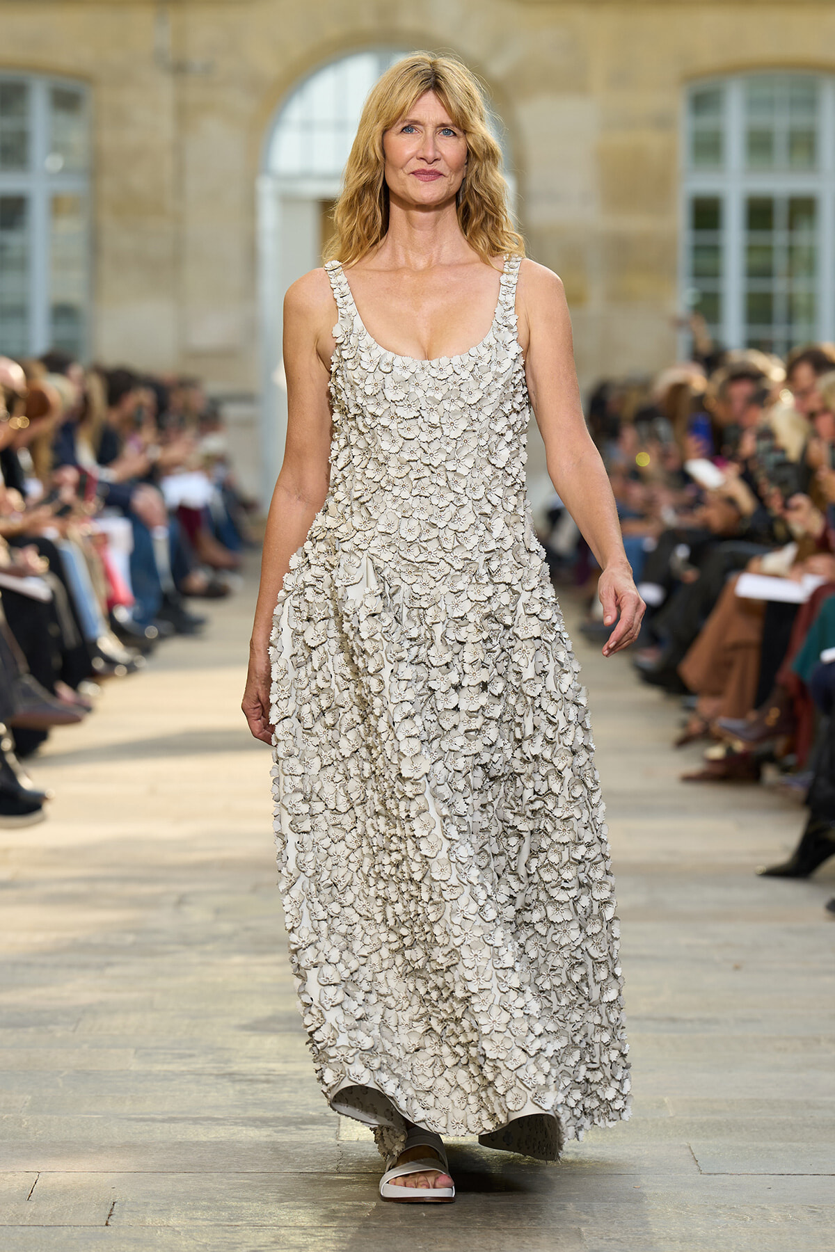 Woman wearing a long cream floral-textured dress walking down a fashion show runway.