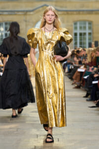 Model walks a fashion runway in a shiny gold metallic dress with puffed sleeves, holding a black handbag and wearing black sandals.
