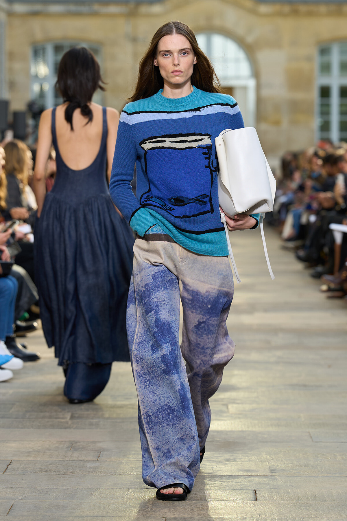 Model walking on a runway outdoors wearing a blue sweater with a mug graphic, light-waded turquoise accents, wide-leg printed pants, and carrying a white bag.