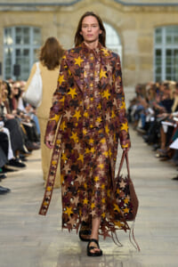 Model walking the runway in a long sheer coat covered in brown, yellow, and beige star shapes, carrying a brown star-patterned tote bag.