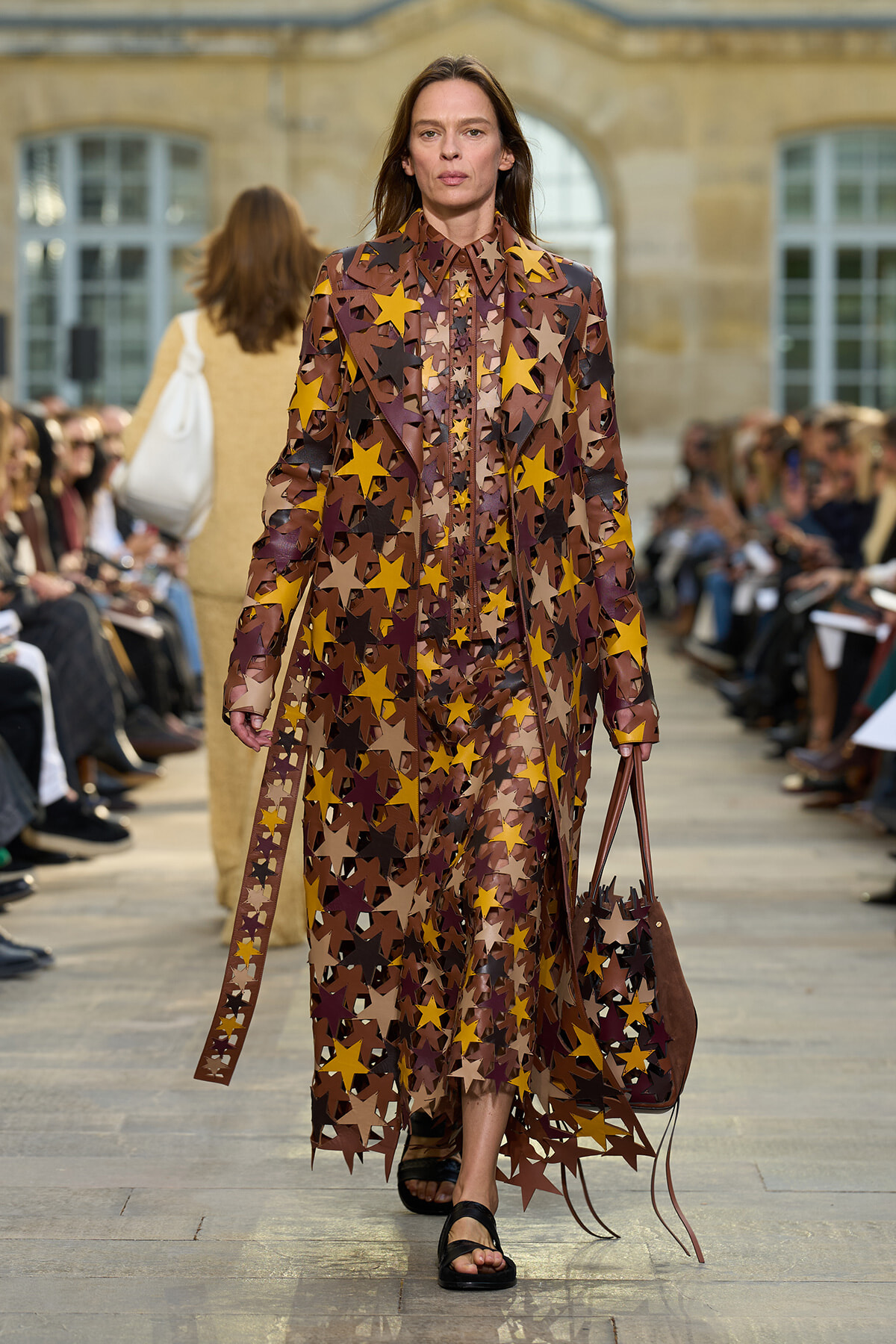 Model walking the runway in a long sheer coat covered in brown, yellow, and beige star shapes, carrying a brown star-patterned tote bag.