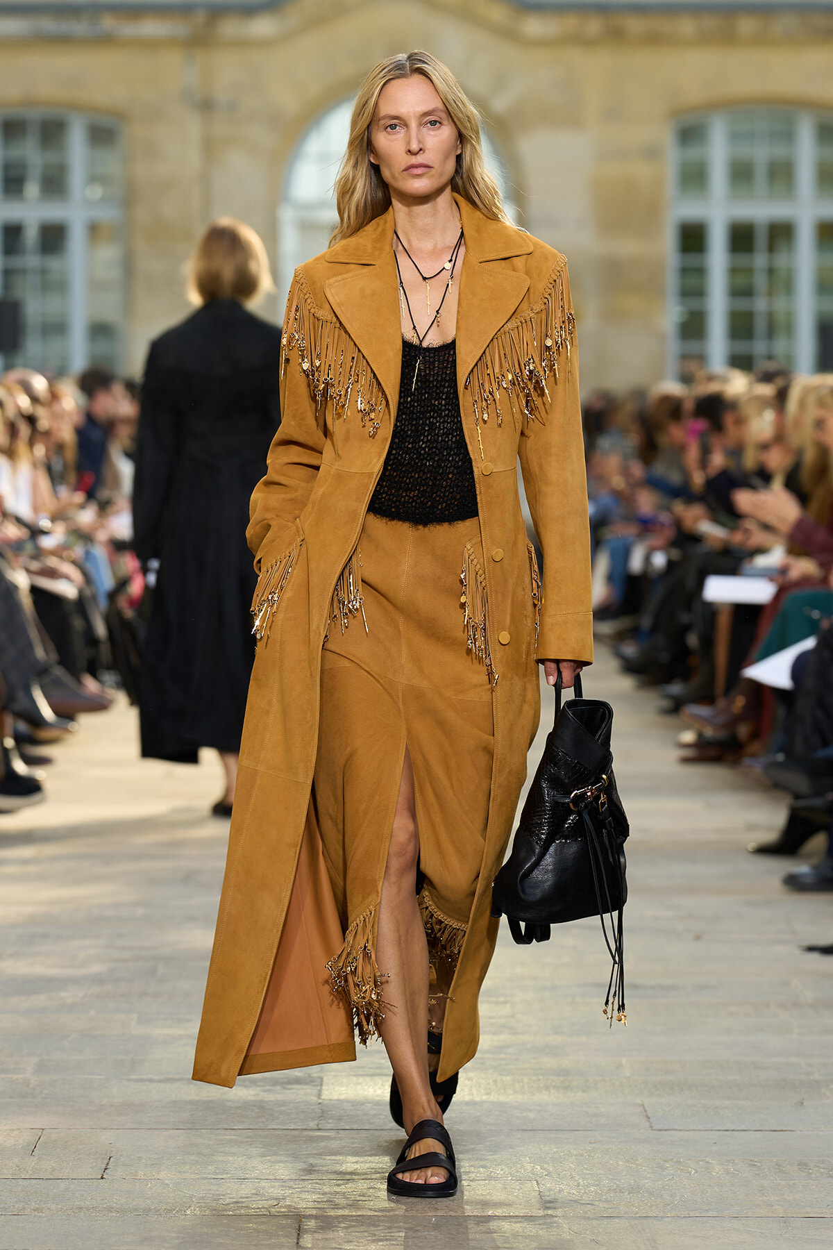 Model walking a runway in a tan suede fringe coat with beaded fringe details, black knit top, and matching skirt with a front slit; carrying a black bucket bag.