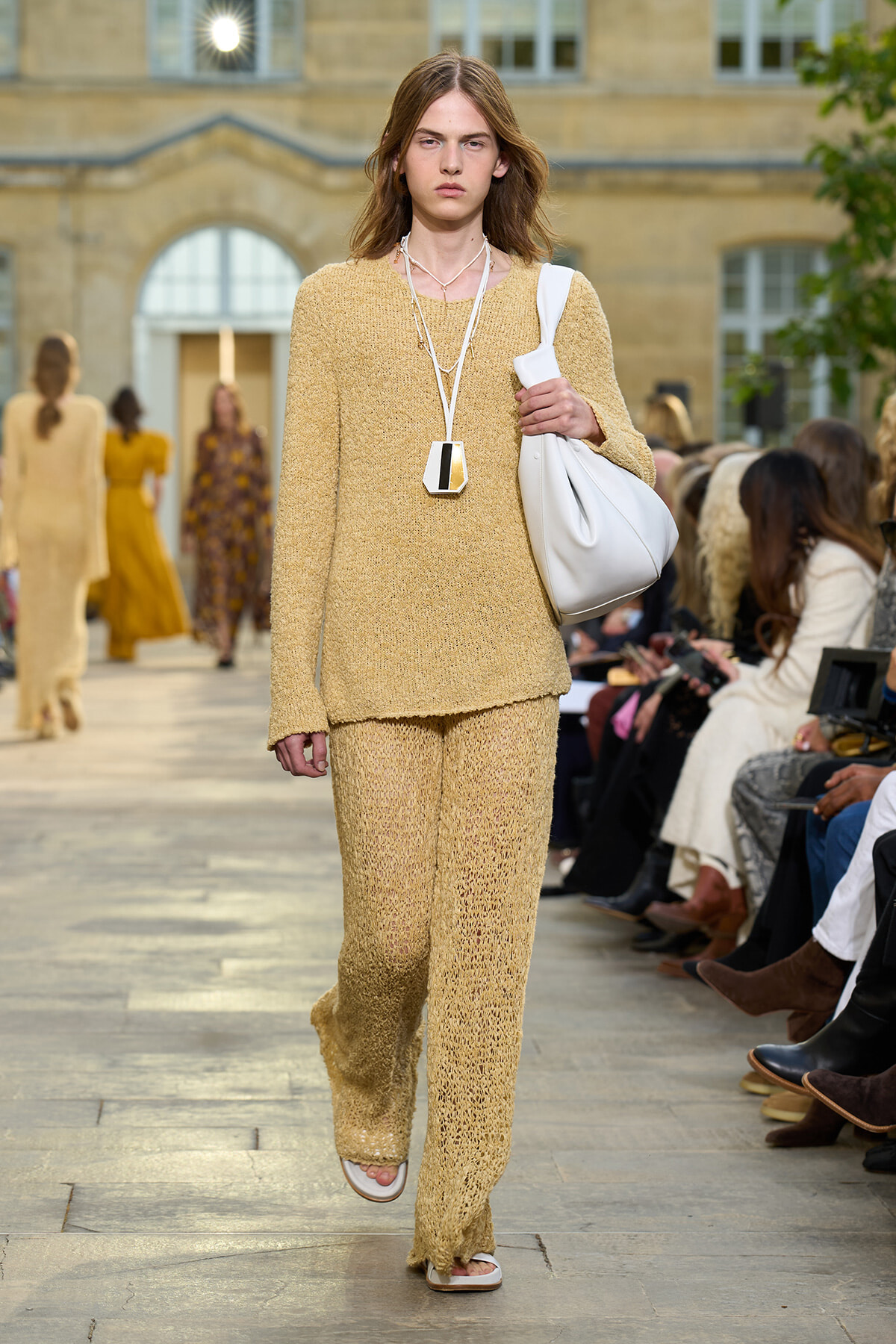 Model walks the runway in a beige knit matching set with a white shoulder bag and geometric pendant necklace at a fashion show.