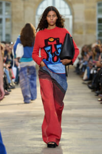 Model walking an outdoor fashion show runway in a red graphic sweater, patchwork jeans, and a black handbag.