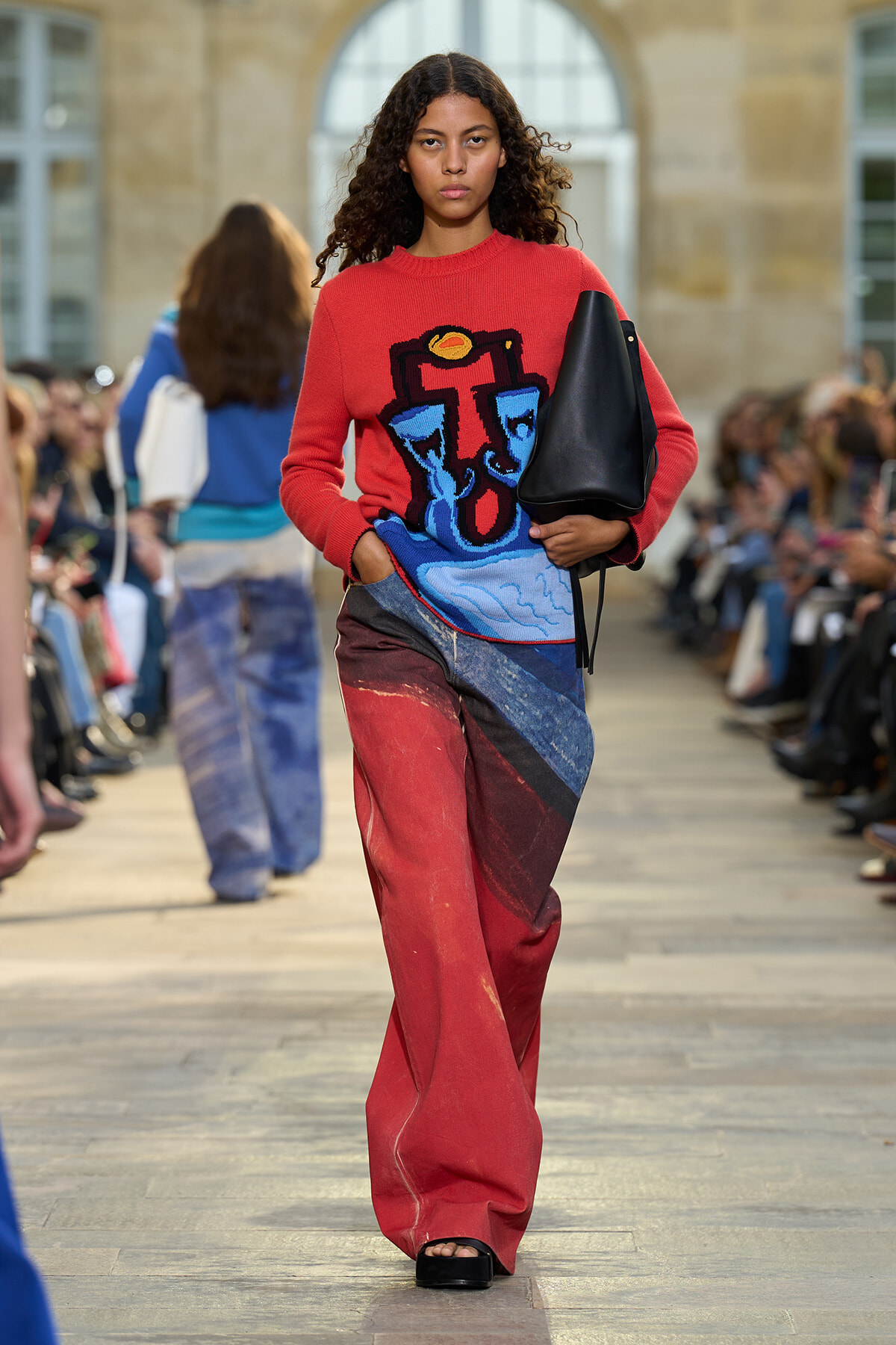 Model walking an outdoor fashion show runway in a red graphic sweater, patchwork jeans, and a black handbag.