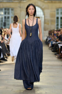 Model walking a fashion runway in a sleeveless dark denim maxi dress with a gathered skirt, wearing layered necklaces and sandals.