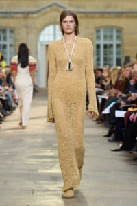 Model walking a fashion runway in a beige crocheted maxi dress with a geometric pendant necklace.