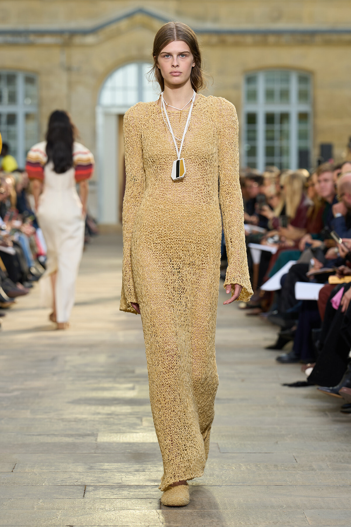 Model walking a fashion runway in a beige crocheted maxi dress with a geometric pendant necklace.