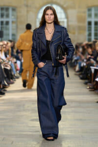 Model walks a runway in a navy leather biker jacket with wide-leg denim trousers, carrying a black handbag.