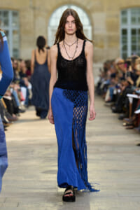 Model walks a runway in a black crochet top and a long blue fringe skirt, with an audience on both sides.