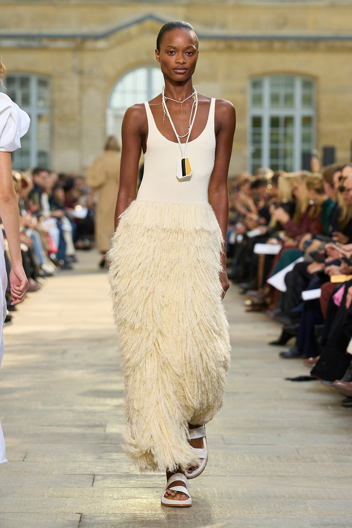 Model wearing a cream sleeveless fringe dress with layered necklaces walking a runway, audience on both sides.