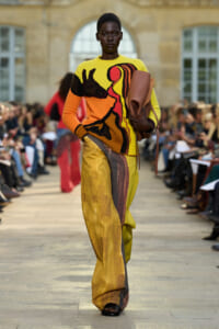 Model walking a fashion show runway in a bright abstract-knit sweater and color-block wide-leg pants, carrying a brown rolled clutch.