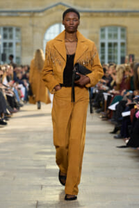 Female model walks the runway in a mustard suede fringe jacket and matching wide-leg pants, holding a black clutch as the audience watches.