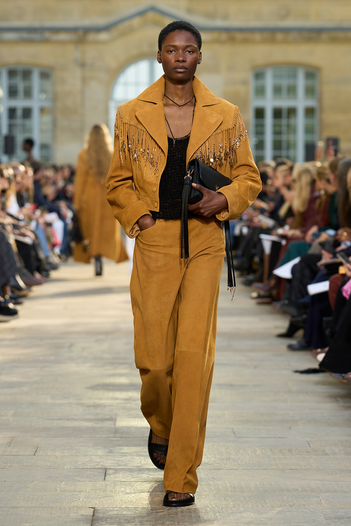 Female model walks the runway in a mustard suede fringe jacket and matching wide-leg pants, holding a black clutch as the audience watches.
