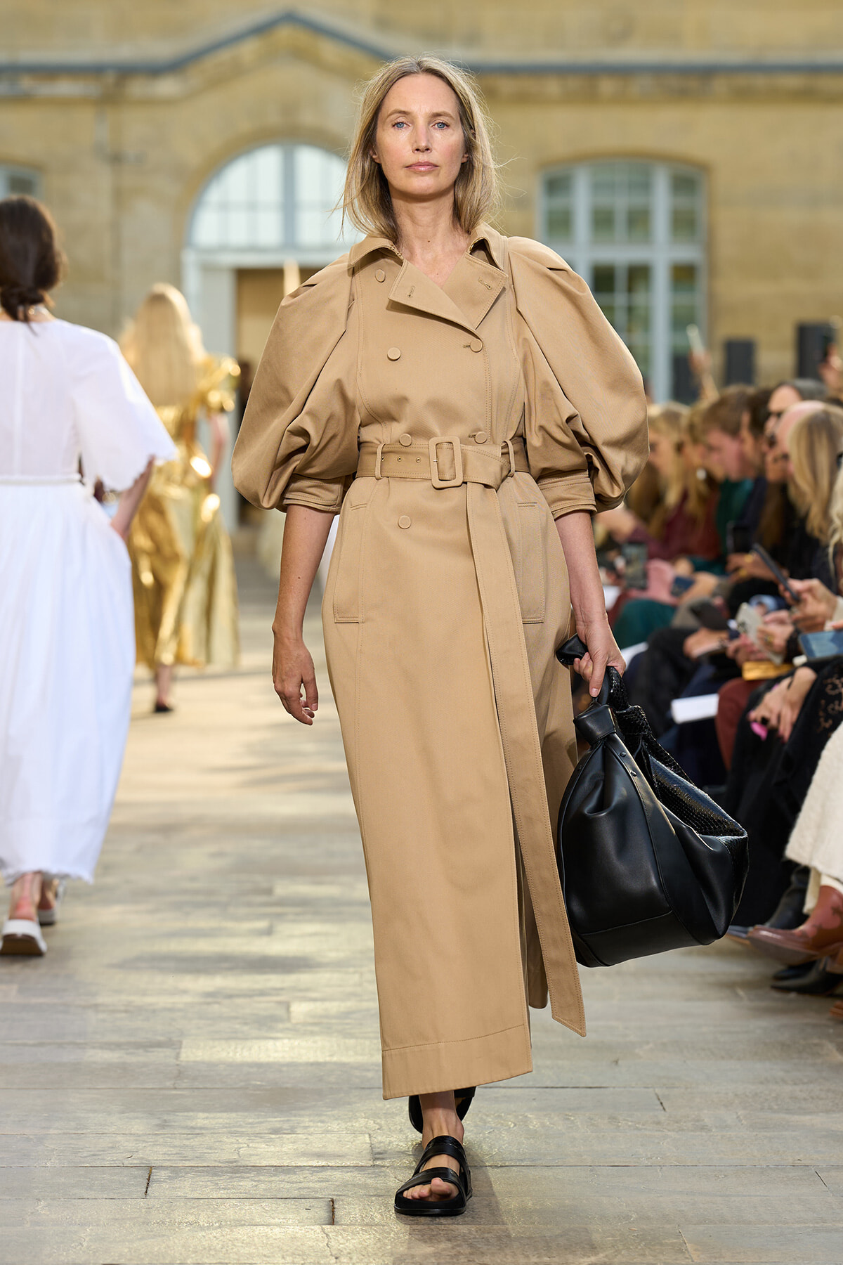 Model marching a runway in a tan trench dress with oversized sleeves and a matching belt, carrying a black handbag.