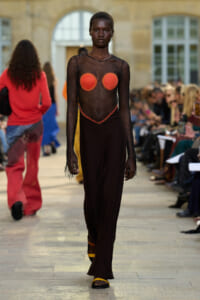 Model walks a runway in a sheer black mesh top with red circular pasties and a long dark skirt, with an audience seated along the catwalk and orange sandals visible.