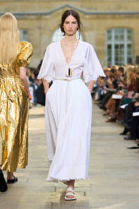Model walks a white maxi dress with fringed sleeves and a rope belt at an outdoor fashion show.