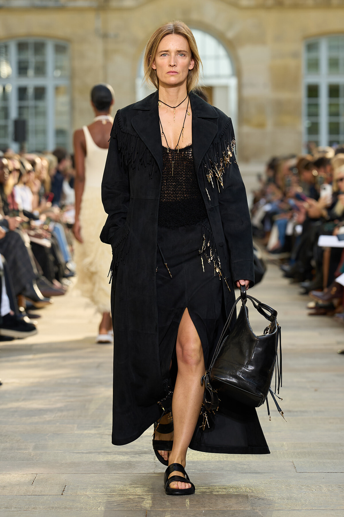 Model walks the runway in a long black fringed coat, black top, and carries a large black handbag amid a fashion show audience.