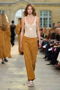 Model walking a runway in a cream crochet tank top and camel wide-leg pants, layered necklaces, and white sandals.