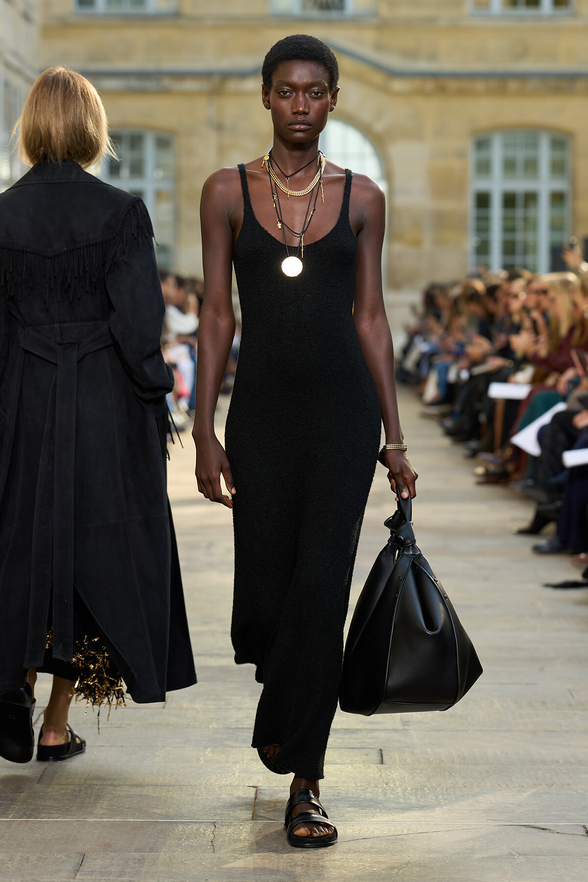 Model walking a fashion runway in a black knit slip dress with layered gold necklaces and a large black handbag.
