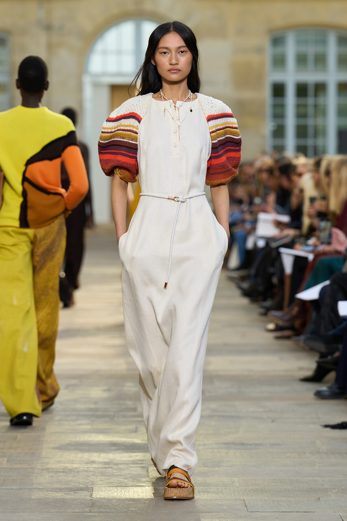 Model walking a runway in a cream jumpsuit with multicolored striped puff sleeves and a belt, audience on both sides.