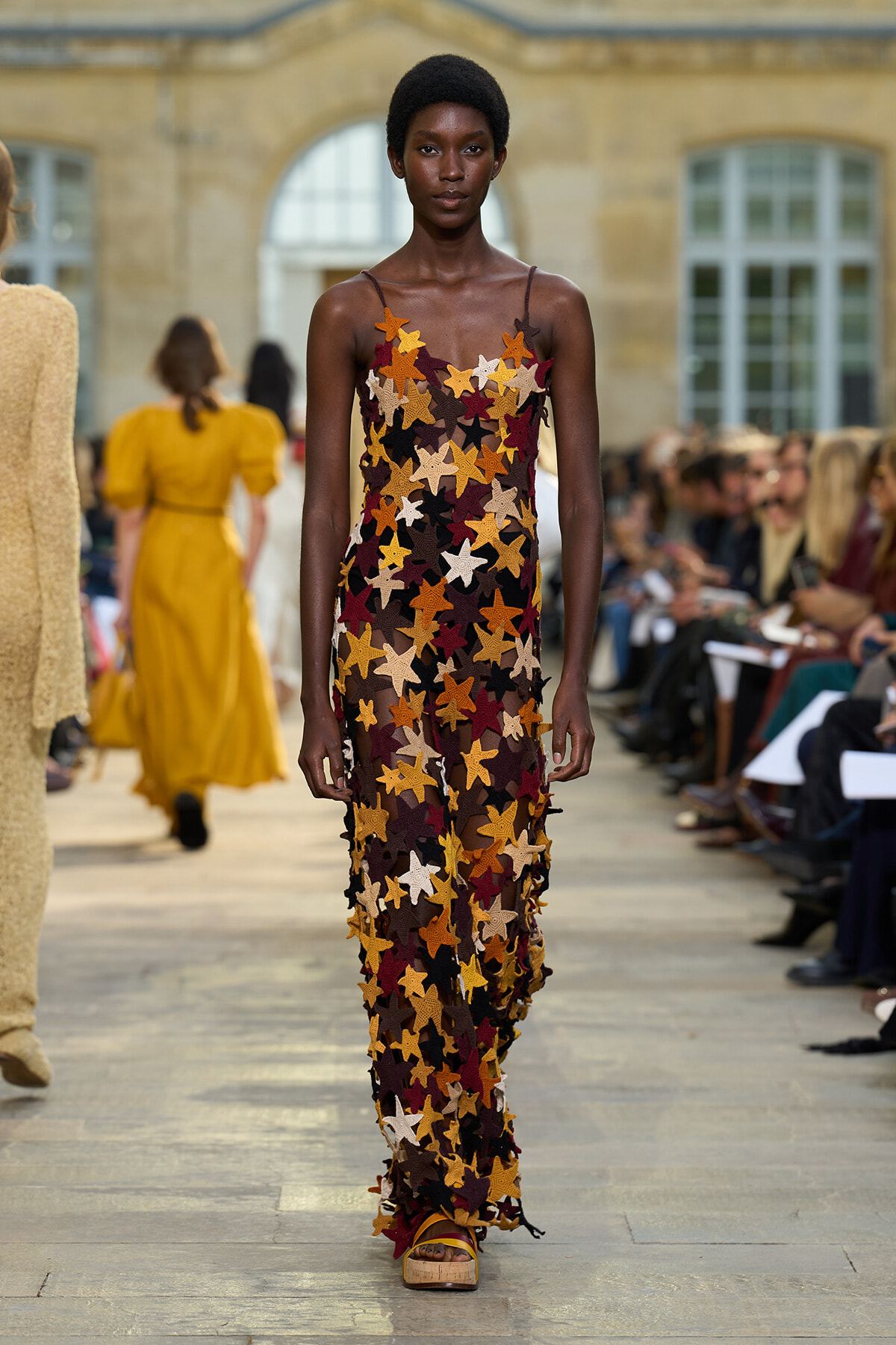 Model walks the runway in a sleeveless, sheer gown covered with multicolored star appliqués (orange, brown, cream, black) during a fashion show, with audience along the catwalk in the background.