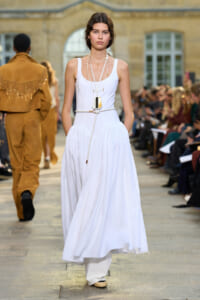 Model walks a runway wearing a white sleeveless maxi dress with layered necklaces and a rope belt, audience on both sides in a fashion show setting.