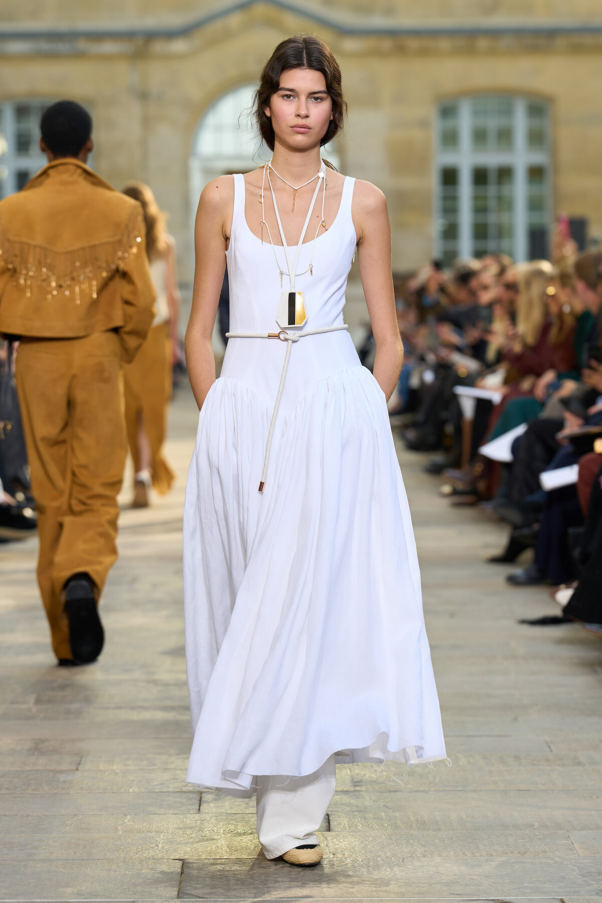 Model walks a runway wearing a white sleeveless maxi dress with layered necklaces and a rope belt, audience on both sides in a fashion show setting.