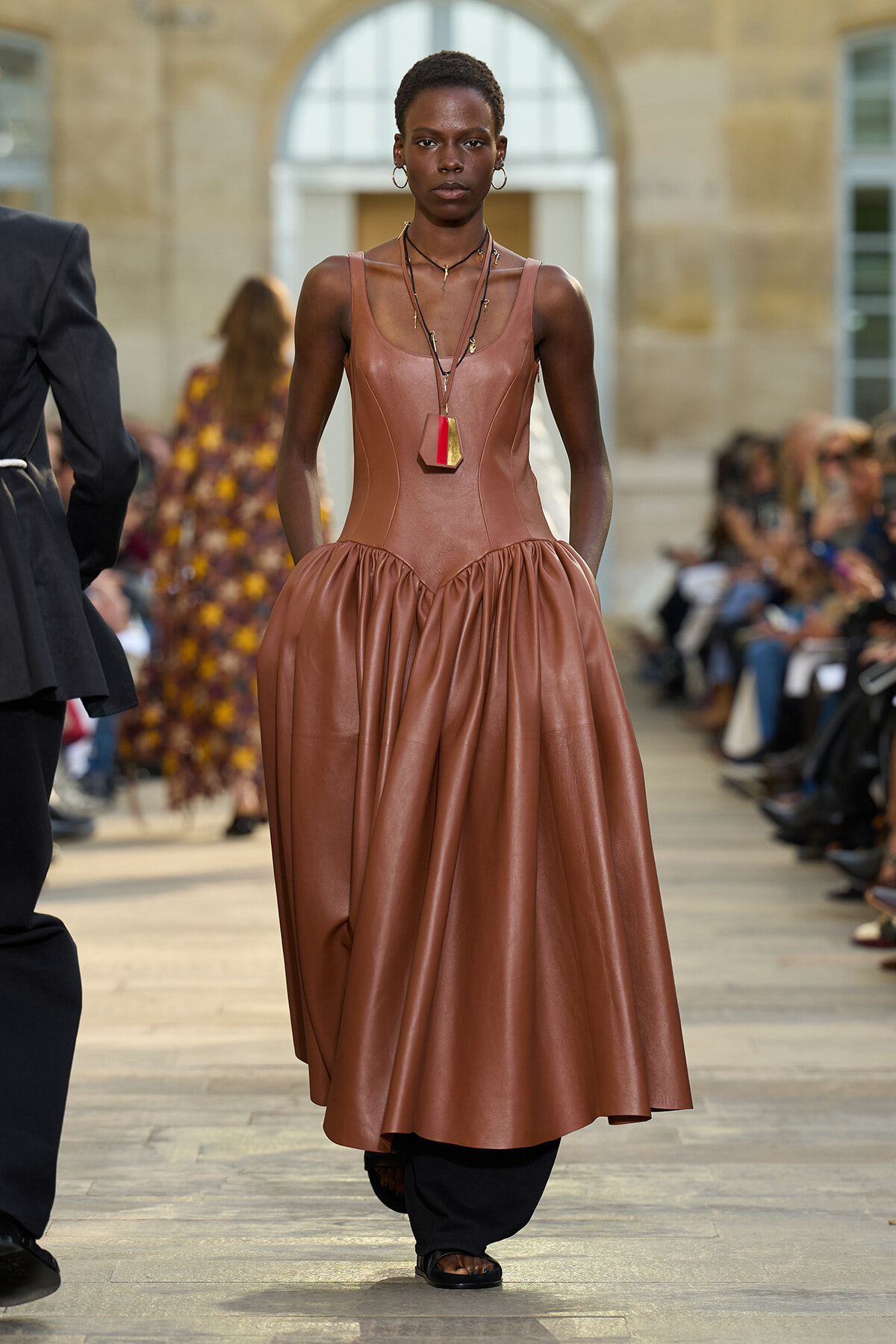 Model walks the runway in a copper-brown leather sleeveless dress with a voluminous skirt and layered necklaces.