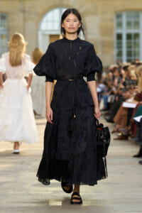 Model wearing a black embroidered dress with puff sleeves walking the outdoor runway at a fashion show.