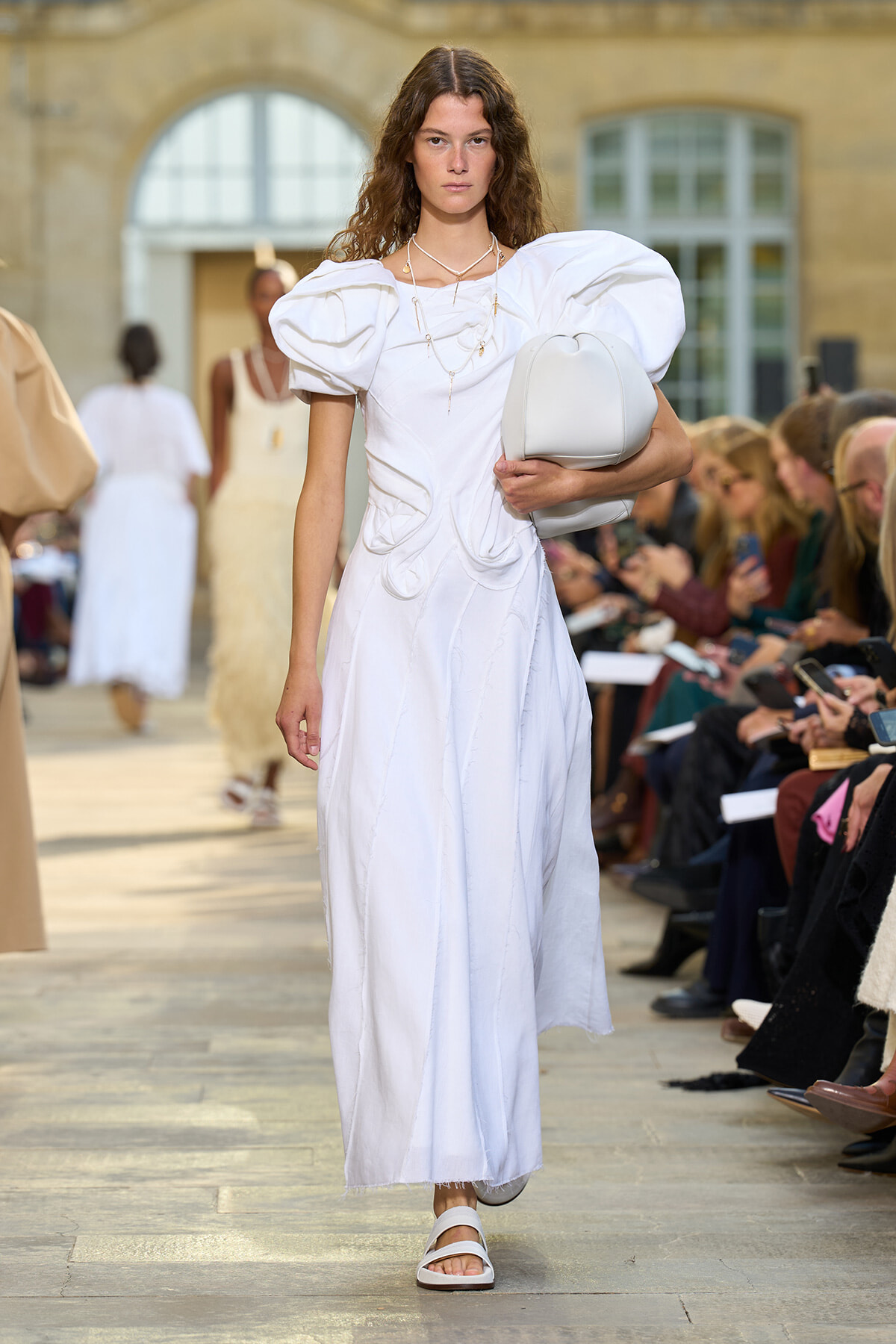 Model walks a white sculptural maxi dress down a runway, carrying a light gray handbag and wearing white sandals, audience watching on sides.