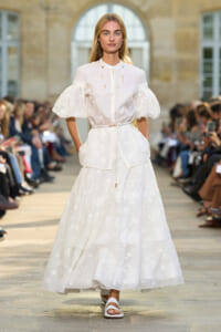 Female model walks the runway in a white embroidered dress with puff sleeves and a slim belt, during an outdoor fashion show.