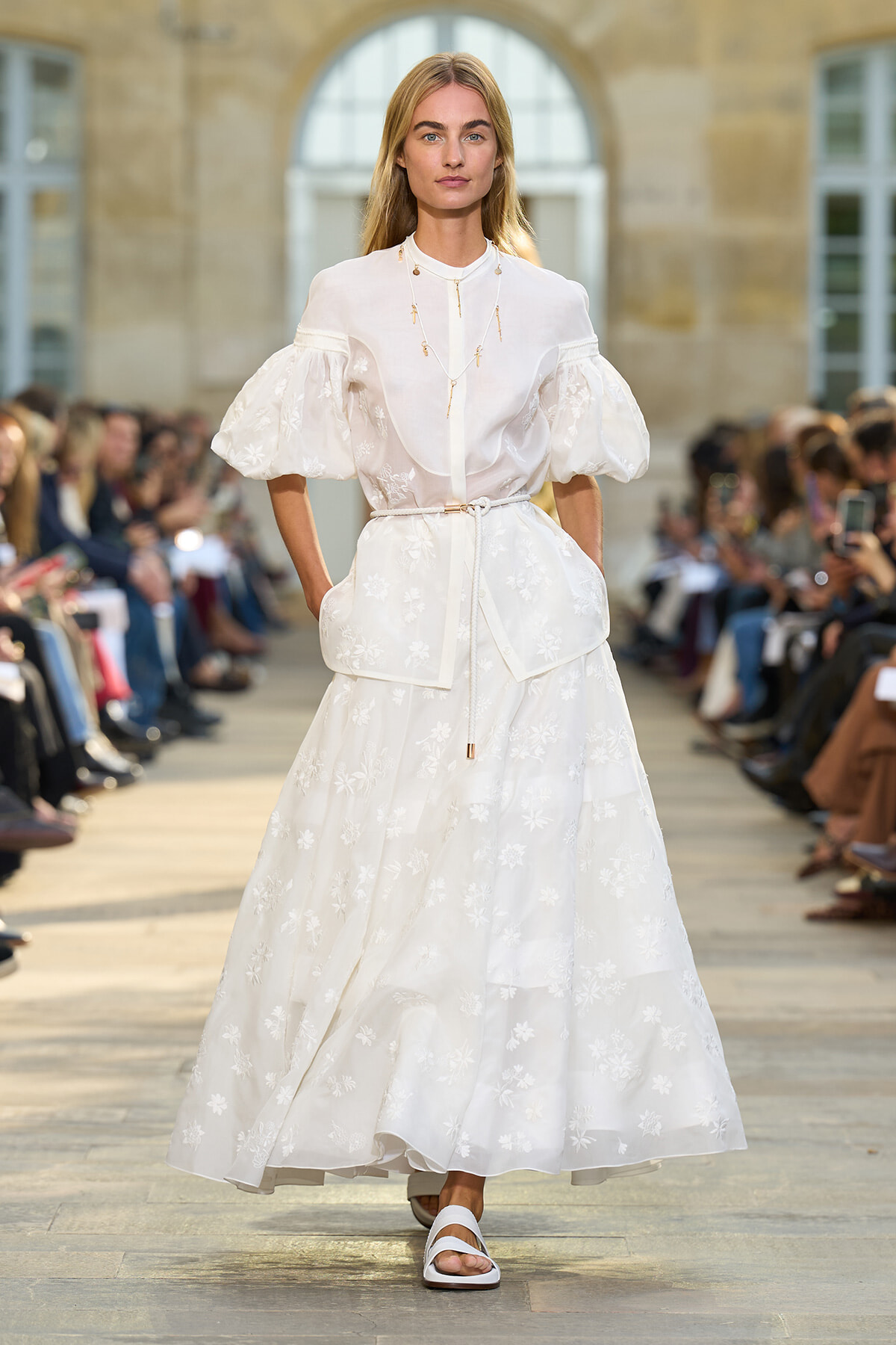 Female model walks the runway in a white embroidered dress with puff sleeves and a slim belt, during an outdoor fashion show.