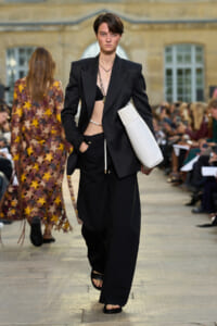 Runway model in a tailored black suit with an open blazer, bralette underneath, and a white oversized clutch at her side.