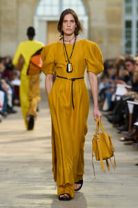 Model walks a fashion runway in a mustard yellow belted dress, carrying a matching yellow handbag; layered necklaces complete the look.