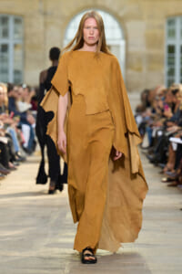 Model walking a runway in a tan suede cape and matching wide-leg outfit with irregular, cut-out edges.