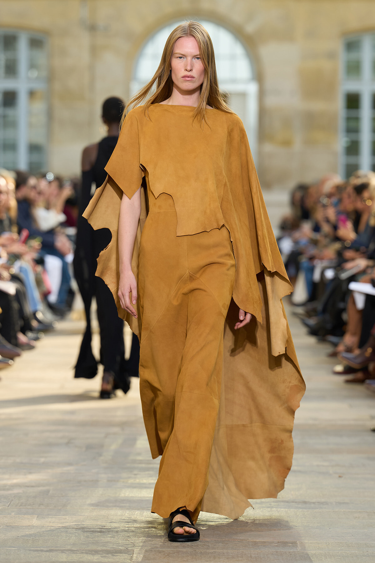 Model walking a runway in a tan suede cape and matching wide-leg outfit with irregular, cut-out edges.