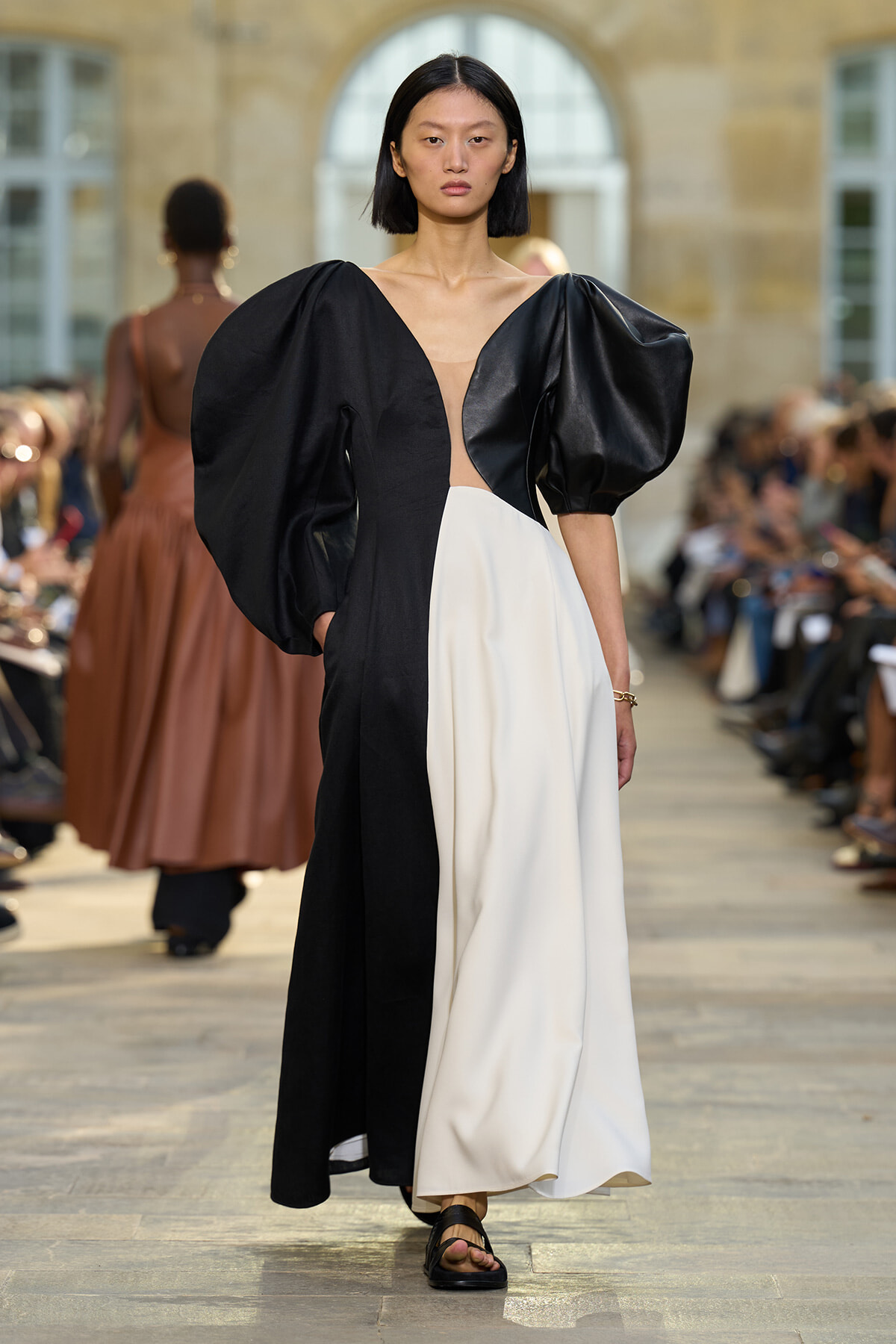 Model walks a runway in a black-and-white color-block gown with oversized puff sleeves and a deep V neckline, audience in background.
