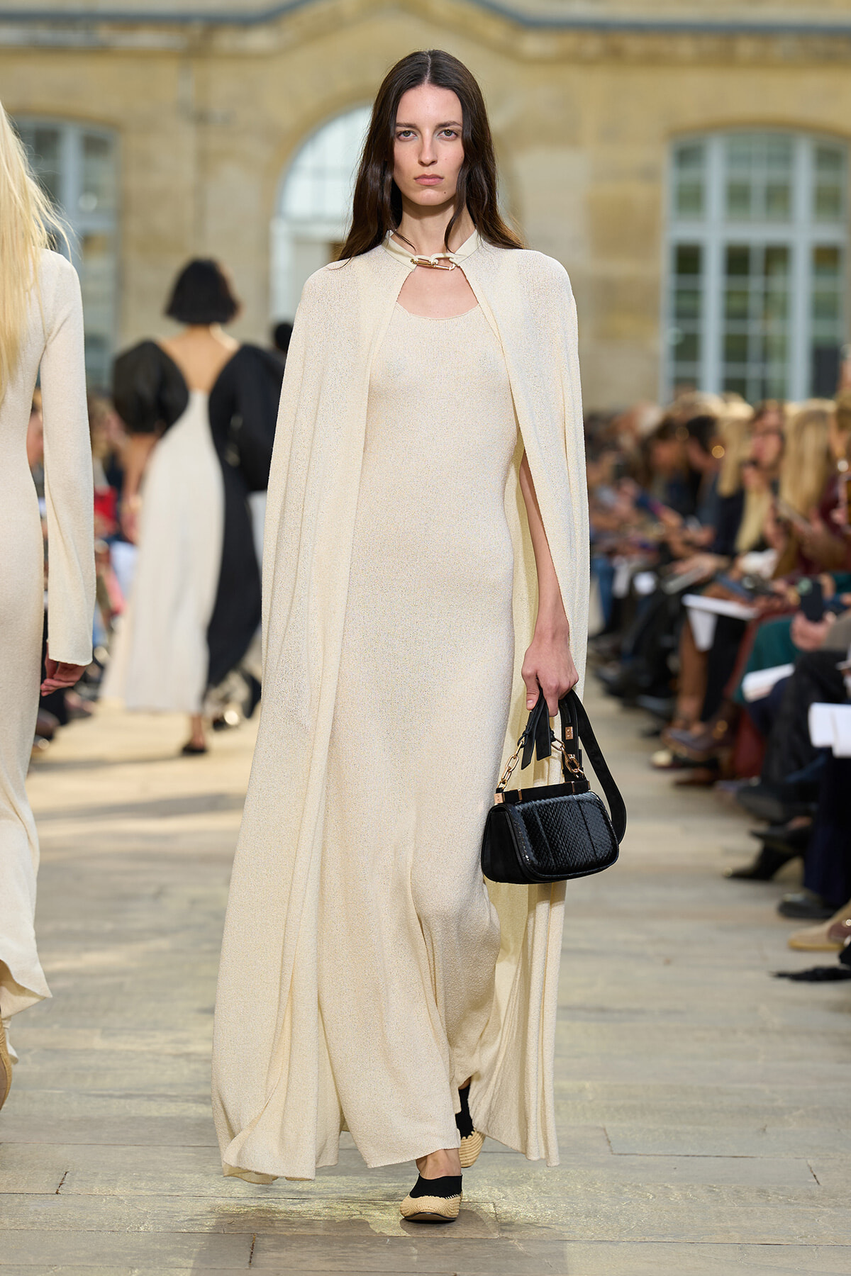 Model walks a runway in a cream-colored, floor-length gown with a matching cape, carrying a small black quilted handbag.