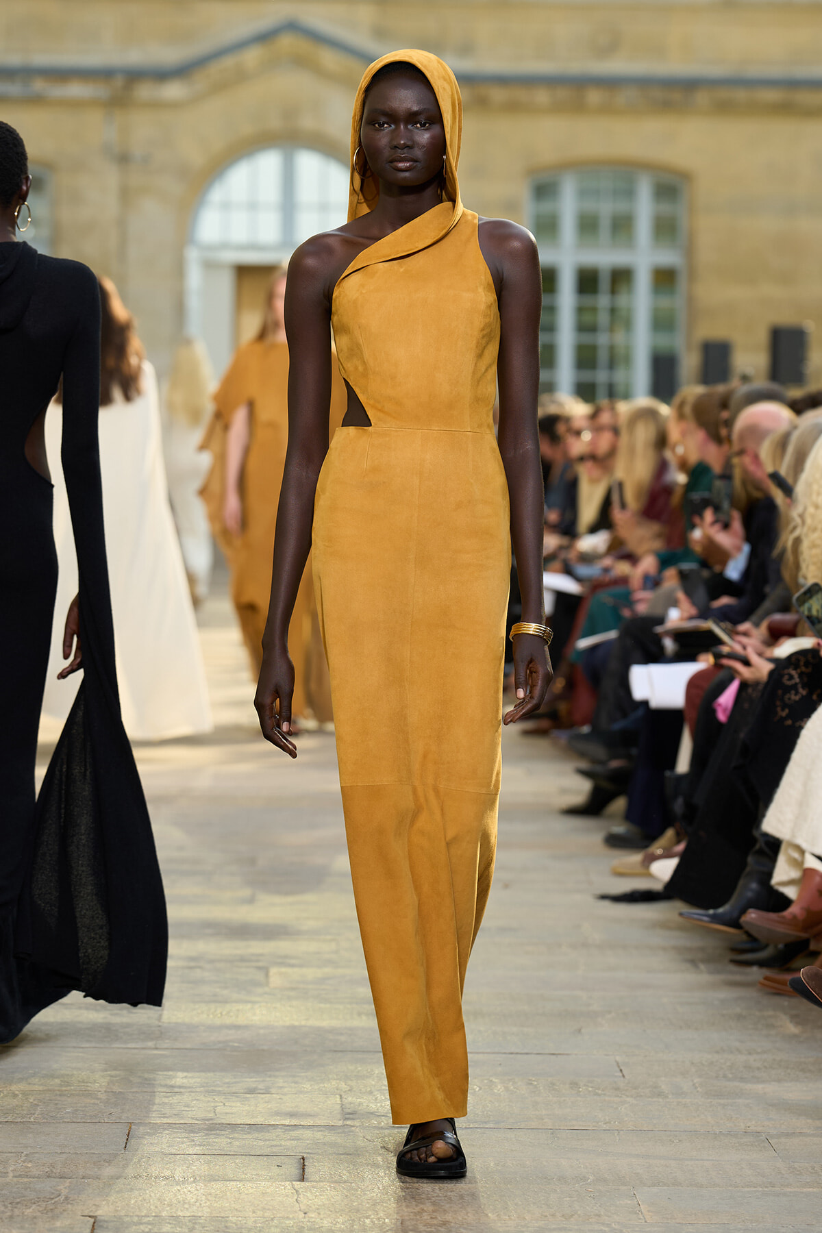 Model on a runway wearing a mustard yellow one-shoulder dress with a hood, strolling an outdoor catwalk with spectators.