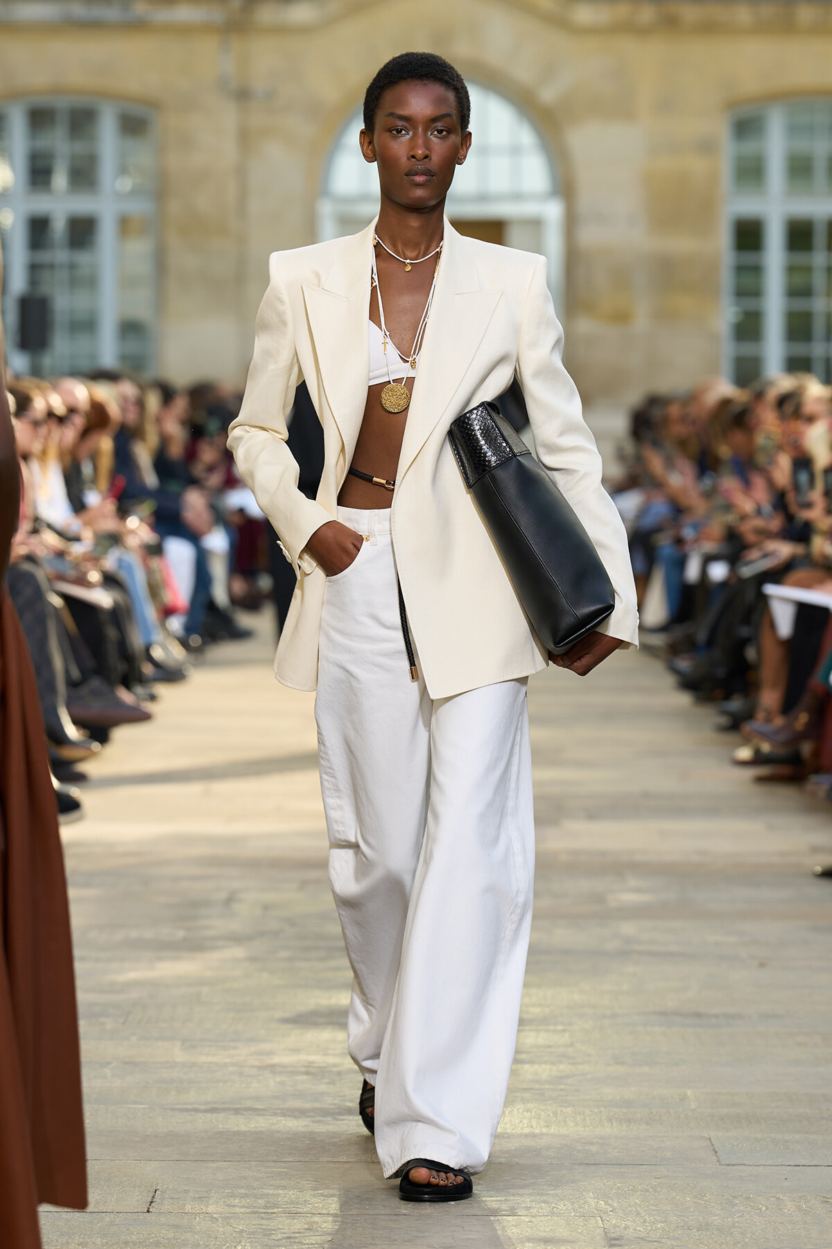 Model walking a fashion runway in a cream oversized blazer and white wide-leg pants, displaying layered gold necklaces and carrying a black clutch.