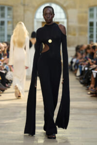 Model walks the runway in a black cut-out jumpsuit with long cape sleeves and gold pendant necklace.