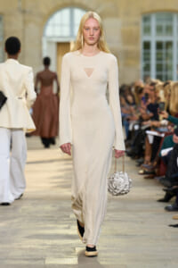 Model walks the runway in a cream knit long-sleeve maxi dress with a small triangle mesh cutout at the chest, holding a textured floral handbag on a chain strap.